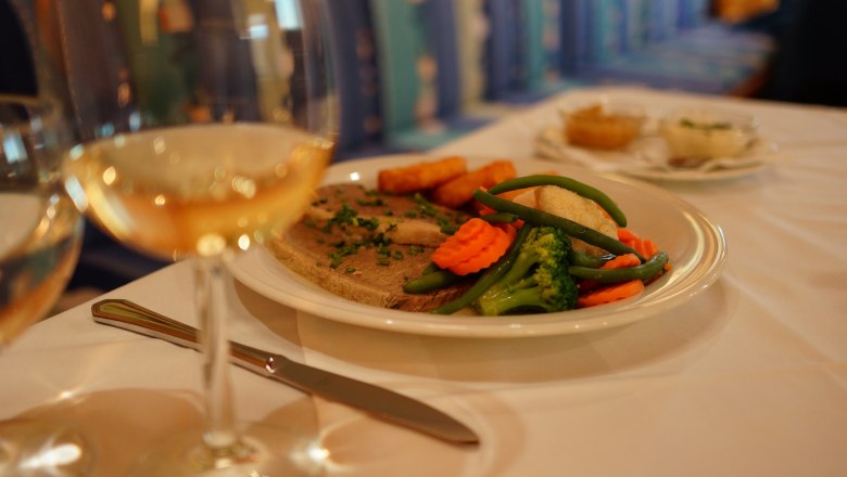 Kulinarik, © Hotel Stich Ein Teller mit Fleisch, Gemüse und Kartoffeln auf einem Tisch, daneben ein Glas Weißwein.