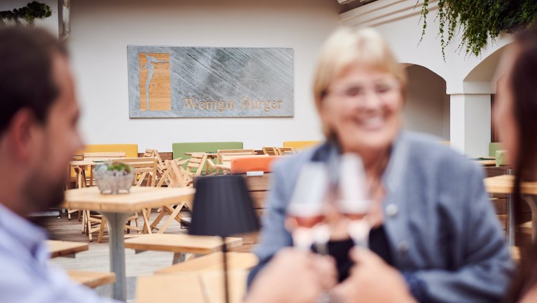 Gemütlicher Innenhof, © Marek Knopp Drei Personen stoßen mit Weingläsern in einem Innenhof an, im Hintergrund ein Schild mit der Aufschrift 'Weingut Burger'.