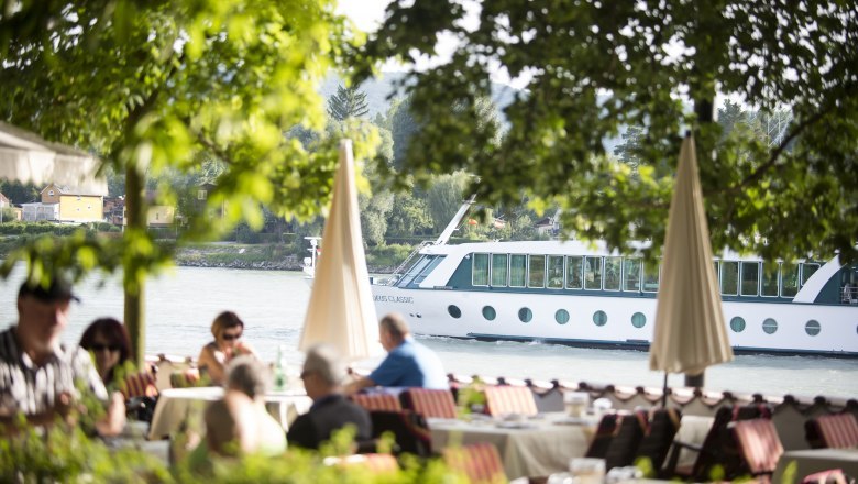 Blick auf die Donau, © Astrid Bartl Ein Flusskreuzfahrtschiff fährt auf der Donau, im Vordergrund sitzen Menschen in einem Café unter Bäumen.