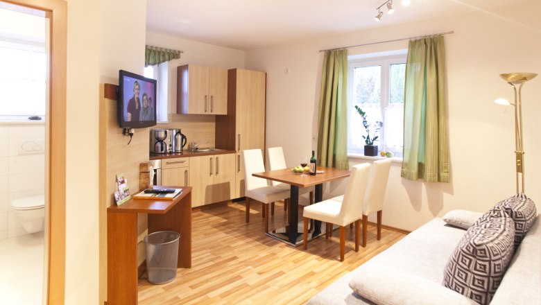 Küche und Aufenthaltsbereich, © Astrid Bartl Moderne Küche mit Esstisch, Sofa und Fernseher in einem hellen Raum.