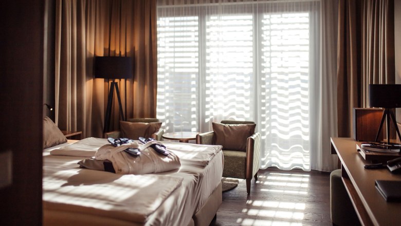 SILENT CLUB rooms at ThermeLaa, © Therme Laa Hotel room with double bed, bathrobes, two armchairs and a table in front of a large window with curtains.