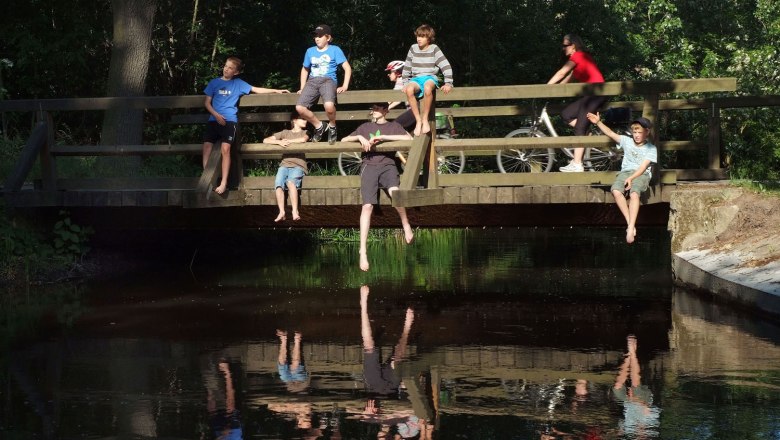 Lindenhof, © Seymannfilm Menschen sitzen auf einer Holzbrücke über einem Fluss, einige lassen die Beine baumeln. Eine Person fährt mit dem Fahrrad über die Brücke.