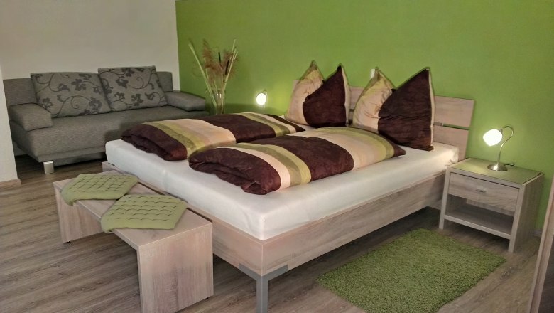 Veltliner Zimmer, © Fam. Wagner Gemütliches Schlafzimmer mit grün gestrichener Wand, Doppelbett, Nachttischlampen und grauem Sofa.