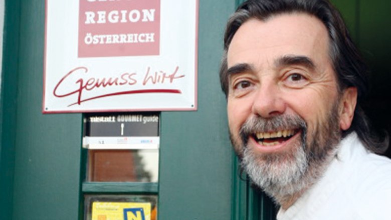 Manfred Buchinger, © Gasthaus Zur Alten Schule Ein Mann mit Bart lächelt vor einem Schild mit der Aufschrift 'Genuss Region Österreich'.