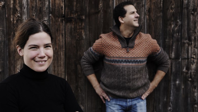 Else & Peter, © Zuschmann-Schöfmann Zwei Personen vor einer Holzwand, eine Frau im Vordergrund lächelt, ein Mann im Hintergrund schaut zur Seite.