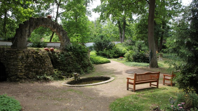 A peaceful garden with a small grotto fountain, surrounded by trees and benches.